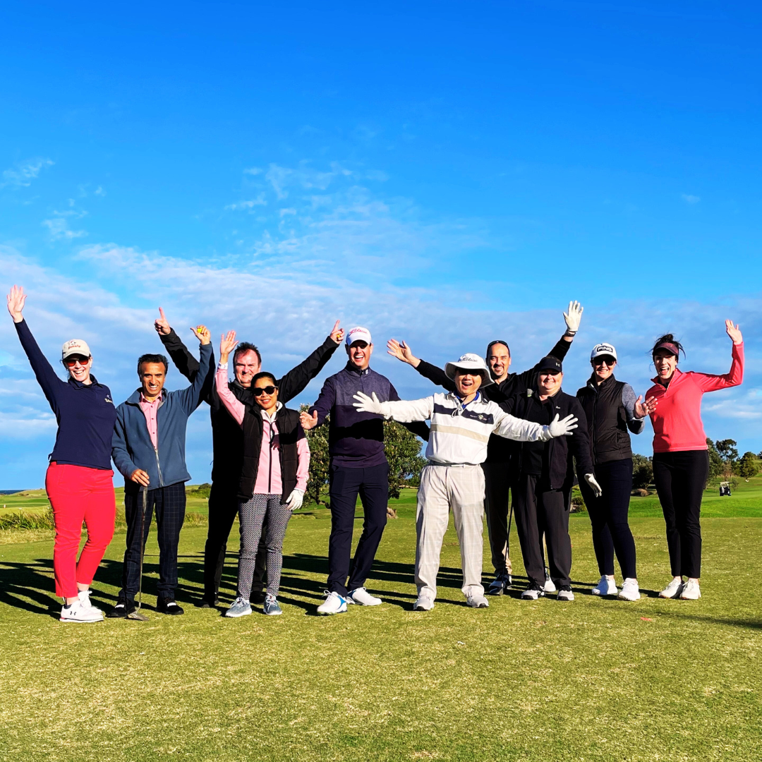 A cheerful group of golfers stand on a sunny fairway with arms raised against a bright blue sky.
